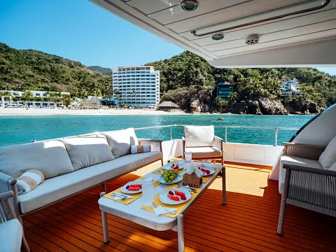 Popa de un yate con sofás y mesa con fruta, frente a una playa con hotel y cerros con selva