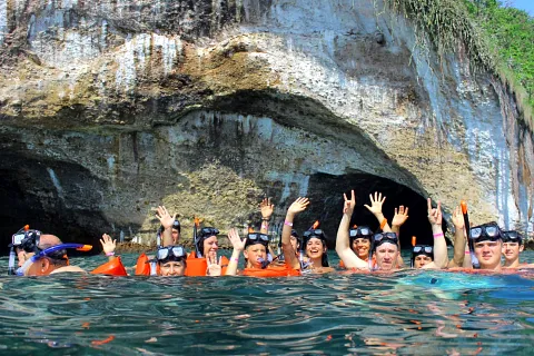 Personas haciendo snorkel saludan frente a la entrada de una cueva en la roca